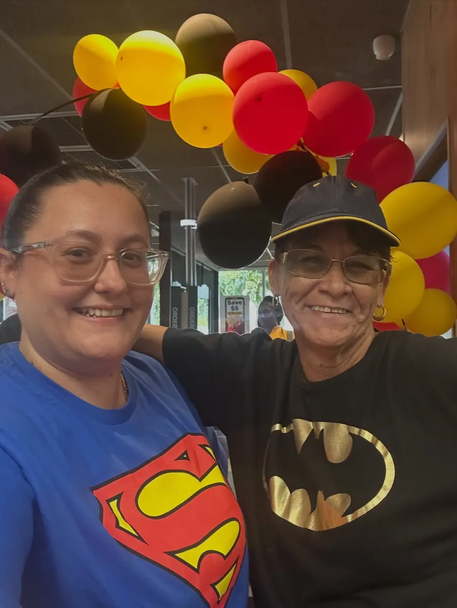 Two crew members in Superman and Batman shirts under a balloon arch